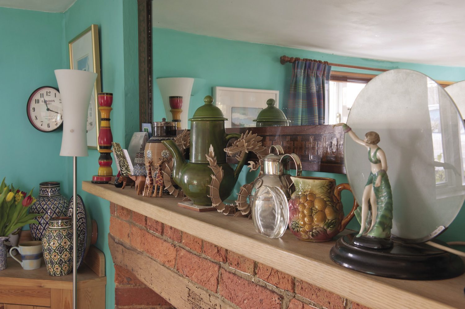 A long mirror with an inlaid wooden frame hangs above the fireplace. The mantelpiece is home to an Arts & Crafts Doulton jug, an olive green lidded pot brought back from a trip to France, a collection of painted wooden African animals and a pair of vibrant candlesticks