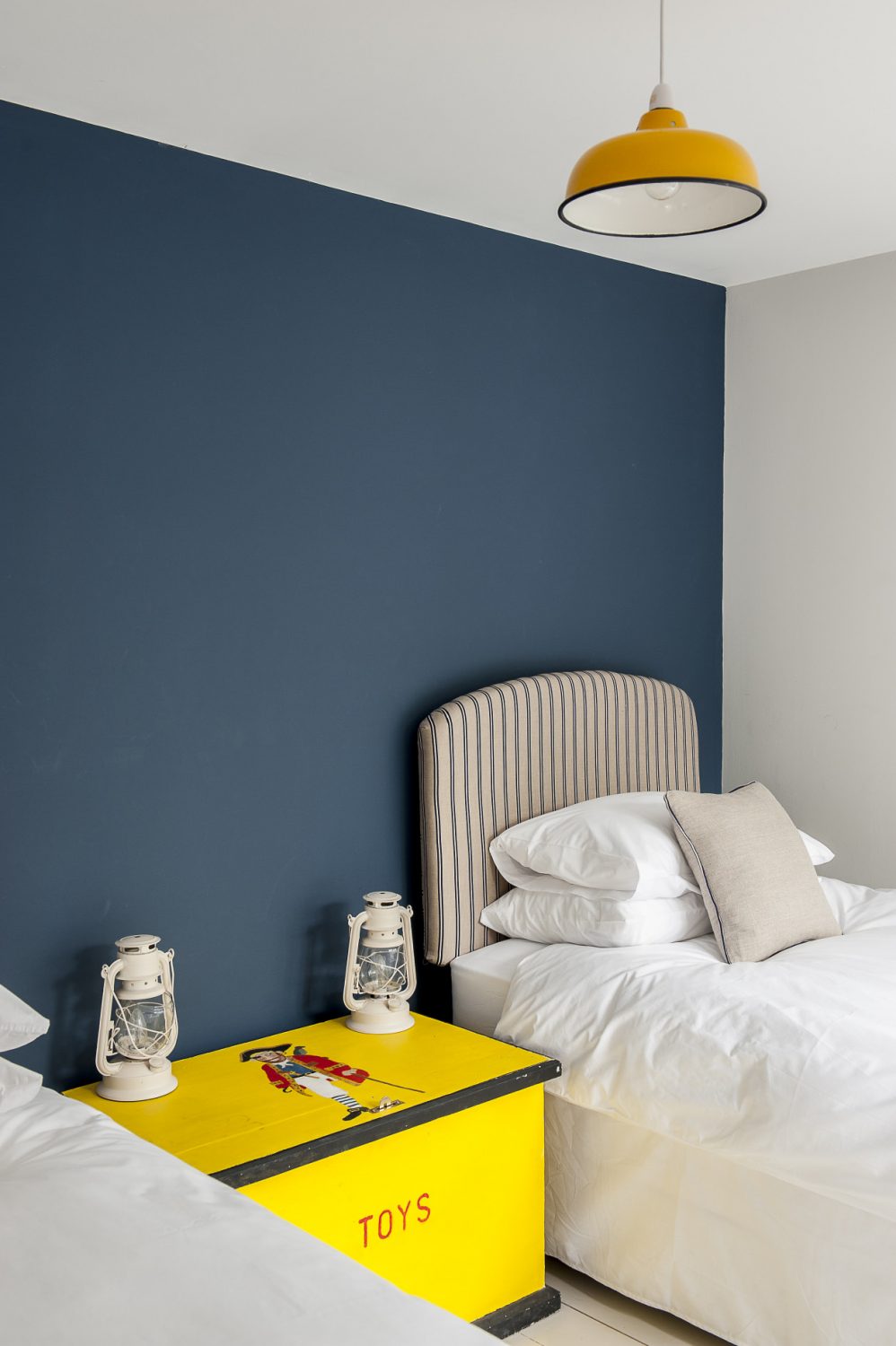 The headboards are Rowan Plowden. Francesca bought the old painted toy box serving as a joint bedside table from a stall in Lydd. The ceiling lampshade is from Hunter Jones. The wall behind the beds is painted in Stiffkey Blue by Farrow & Ball. The hurricane lamps on the chest are from Dunelm