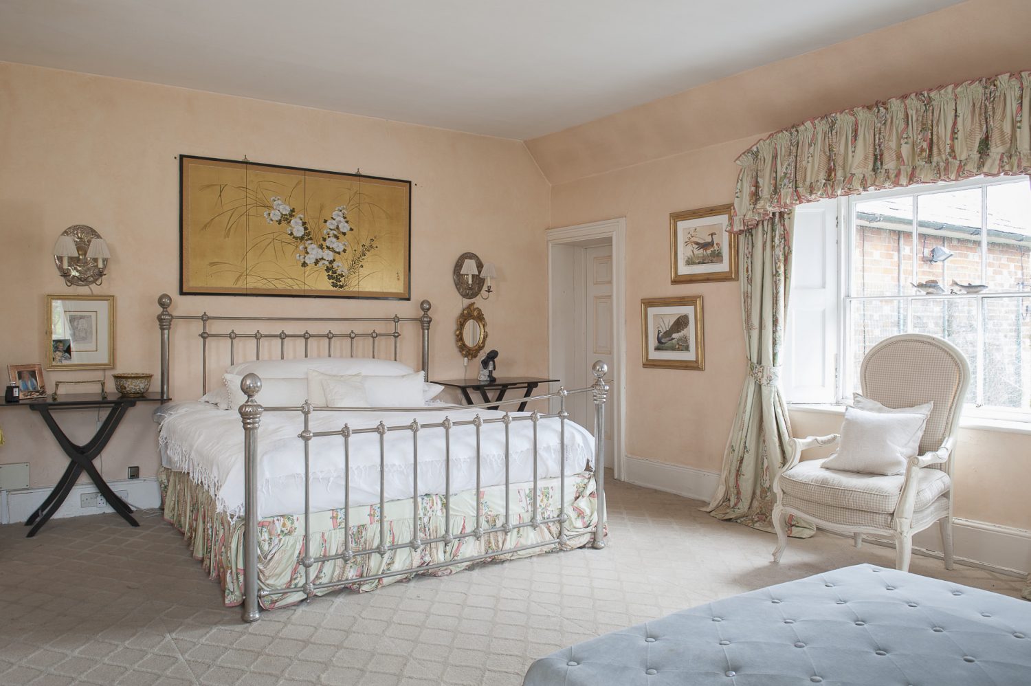 Peachy cream paintwork offsets oriental paintings of birds and flowers, while light dances off the metal bedstead and gilt light sconces. A marble fireplace at the foot of the bed framed on either side by heavy, patterned curtains, gives the room a sense of grandeur as well as softness