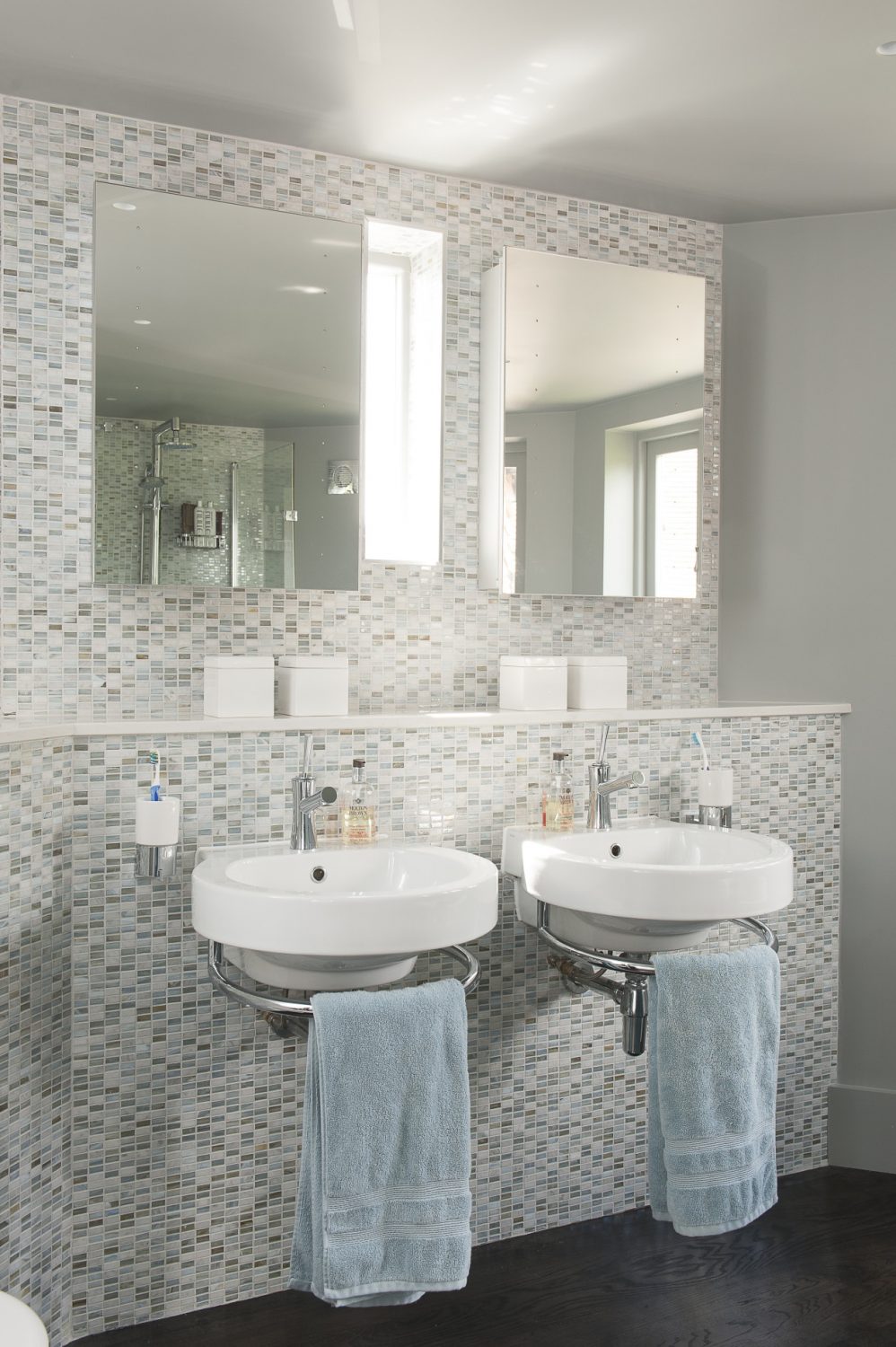 The natural light bounces off the iridescent rectangular tiles covering the wall behind the twin basins