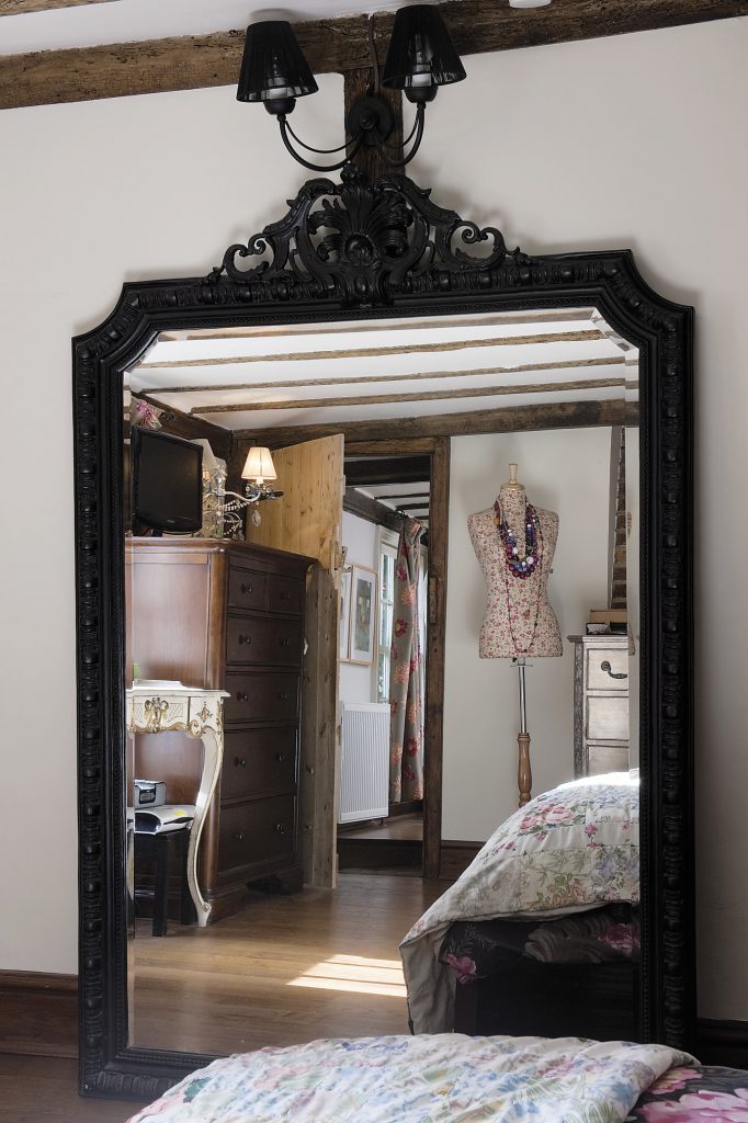The master bedroom. An enormous mirror with an Empire style black frame has been propped up underneath a black wall sconce with finely taut ‘string’ shades A spriggy flower print covers the tailor’s mannequin that displays a collection of vintage and African beads that Louise collected on her travels