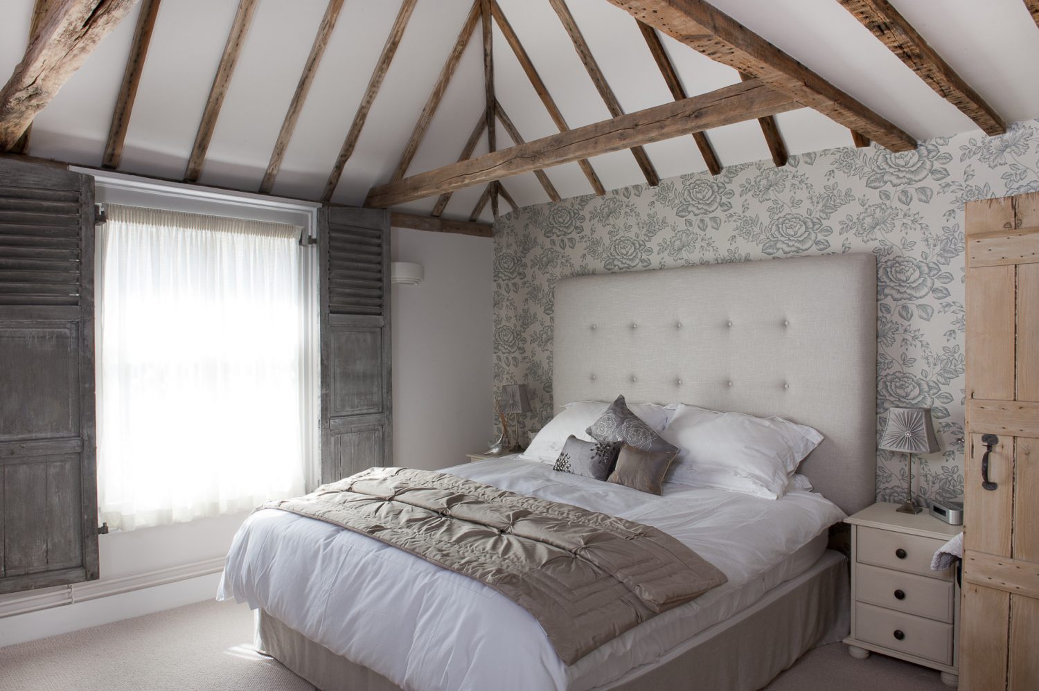 In the glamorous master bedroom a bed dressed with cotton and satin bedclothes is set against a feature wall of Sanderson wallpaper. Plain muslin is draped across the window for privacy during the day and a pair of working shutters with louvred panels can be drawn across at night