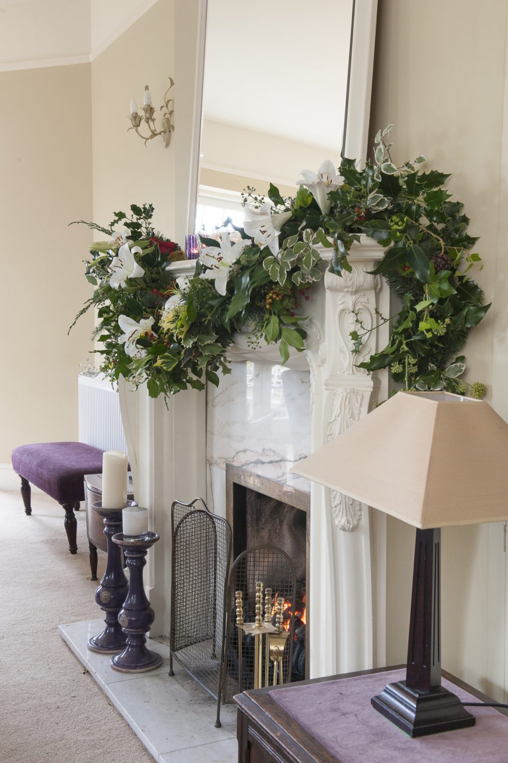 In the drawing room a marble fireplace has been adorned with a swag of berried holly, dark evergreen leaves and white stargazer lilies. The Christmas tree has been decorated with purple and gold ornaments