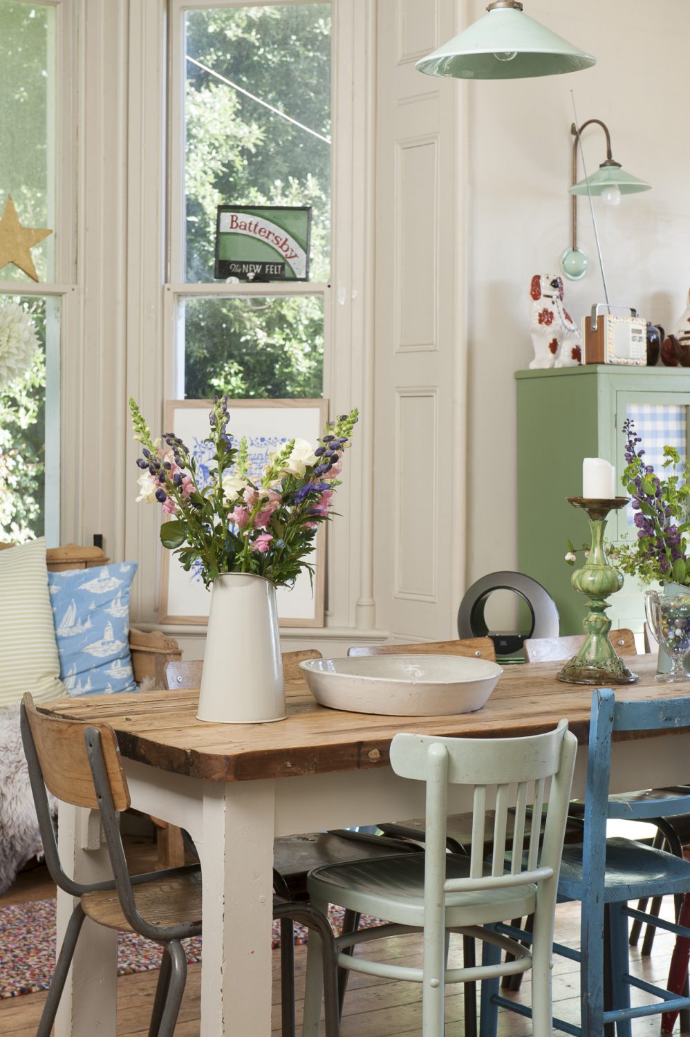 A verdigris coloured French rise and fall pulley light hangs from the very tall ceiling over a long, narrow, scrubbed wooden refectory table that has a collection of vintage school and bentwood chairs around it and a jug of simple English garden flowers at one end