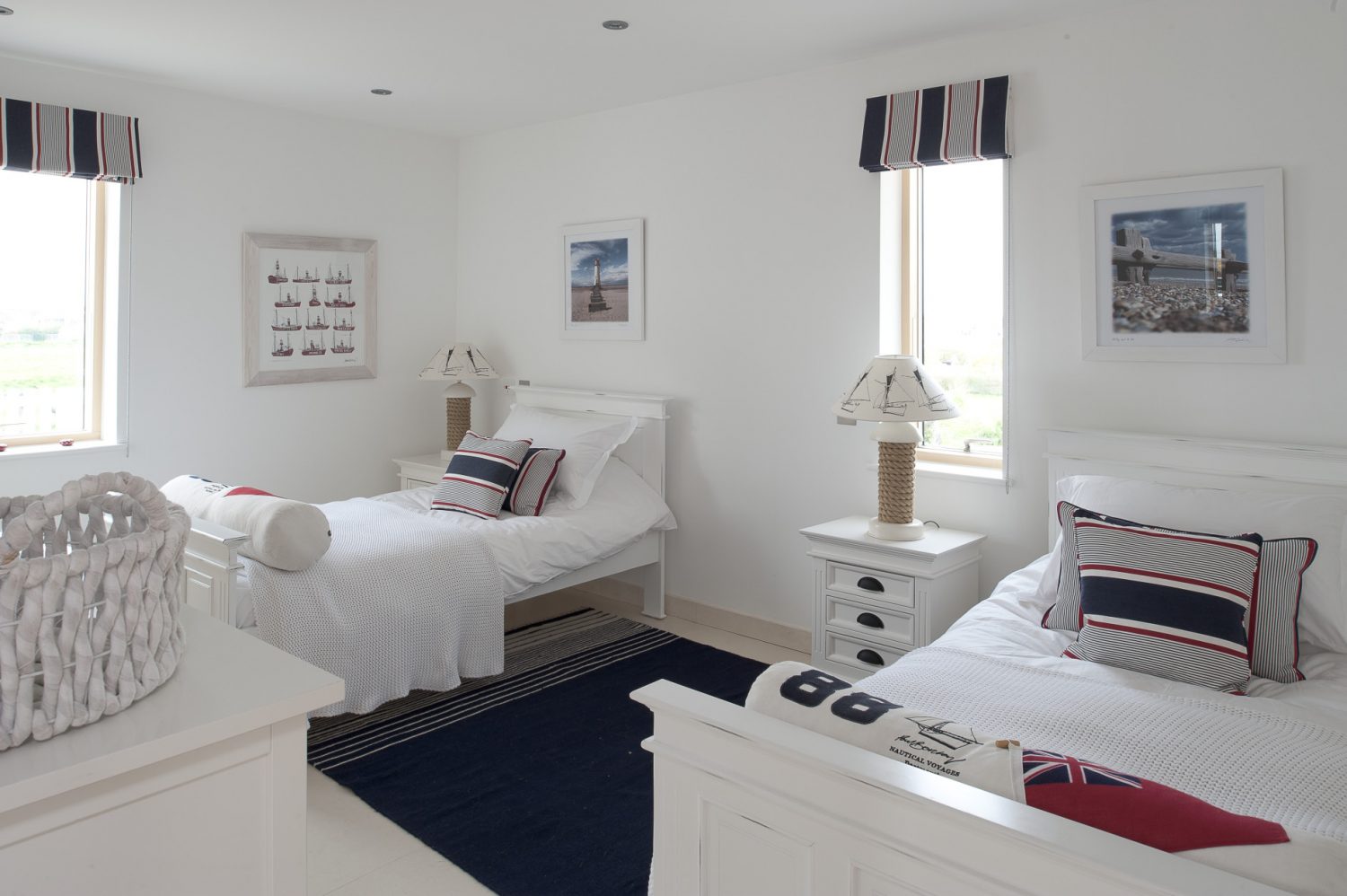 Downstairs, a corridor leads to a twin bedroom with white sleigh beds, knitted covers and red, white and navy blue striped blinds and cushions