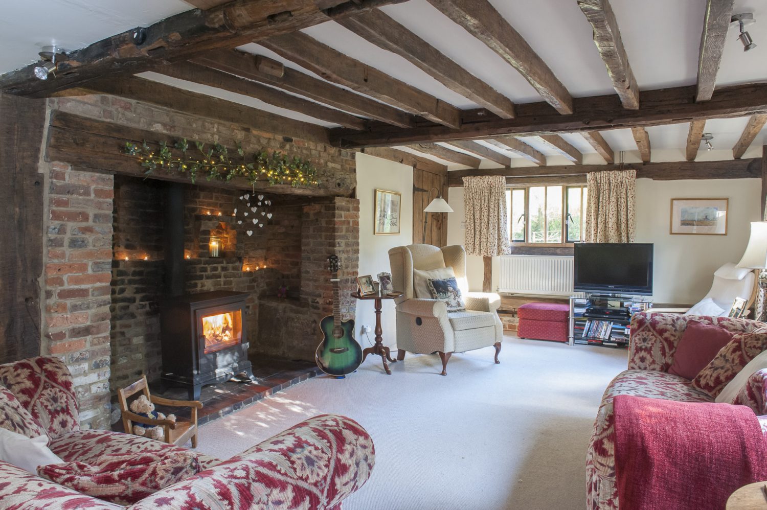 In the sitting room, the second inglenook fireplace shows clearly where the floor has been lowered. Two comfortable tapestry sofas in rich plum tones, complement delicately patterned curtains made by Bell House Fabrics