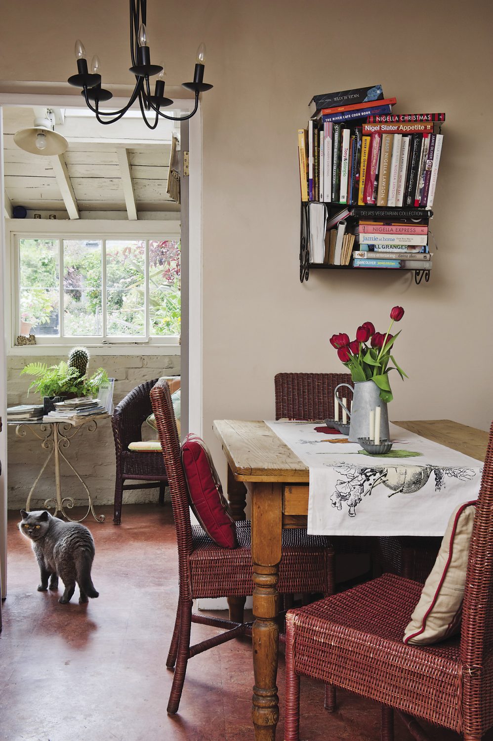 Wicker chairs surround the wooden table in the kitchen area. Above it, metal shelves house Ann’s collection of cookery books
