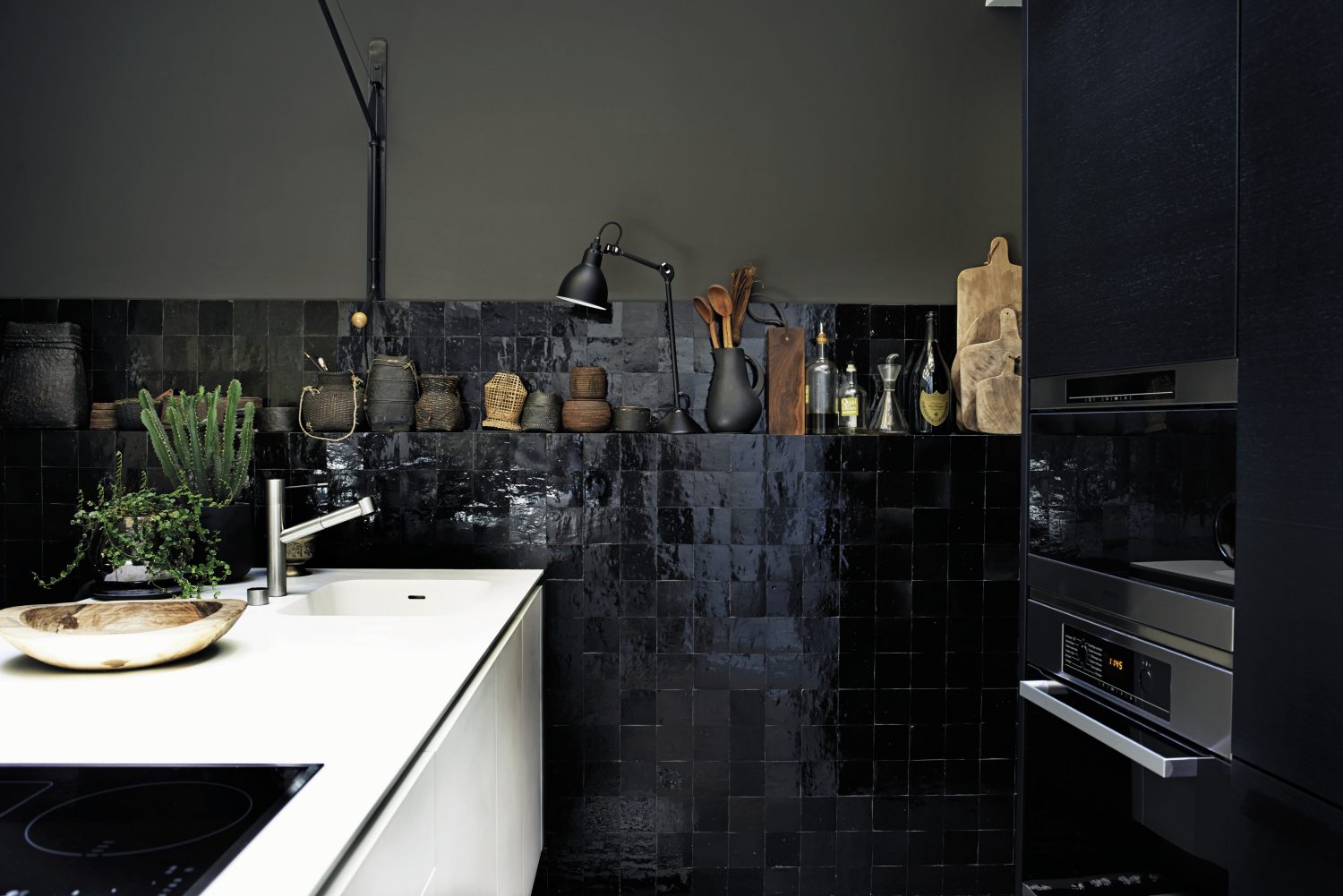 The black and white kitchen is lit by natural light from an atrium, so despite the glossy black tiles and bitter chocolate walls, the space is bright. A white island conceals storage and houses a sink and hob/stovetop. The part-tiled wall is built out enough to include a shallow shelf for utensils, chopping boards, bottles of olive oil, African baskets and a black Anglepoise lamp.