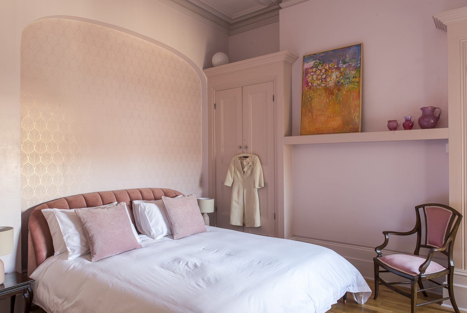 A guest bedroom features a padded and buttoned pink velvet headboard against a wall of deco wallpaper, which gives it a bit of a Biba air