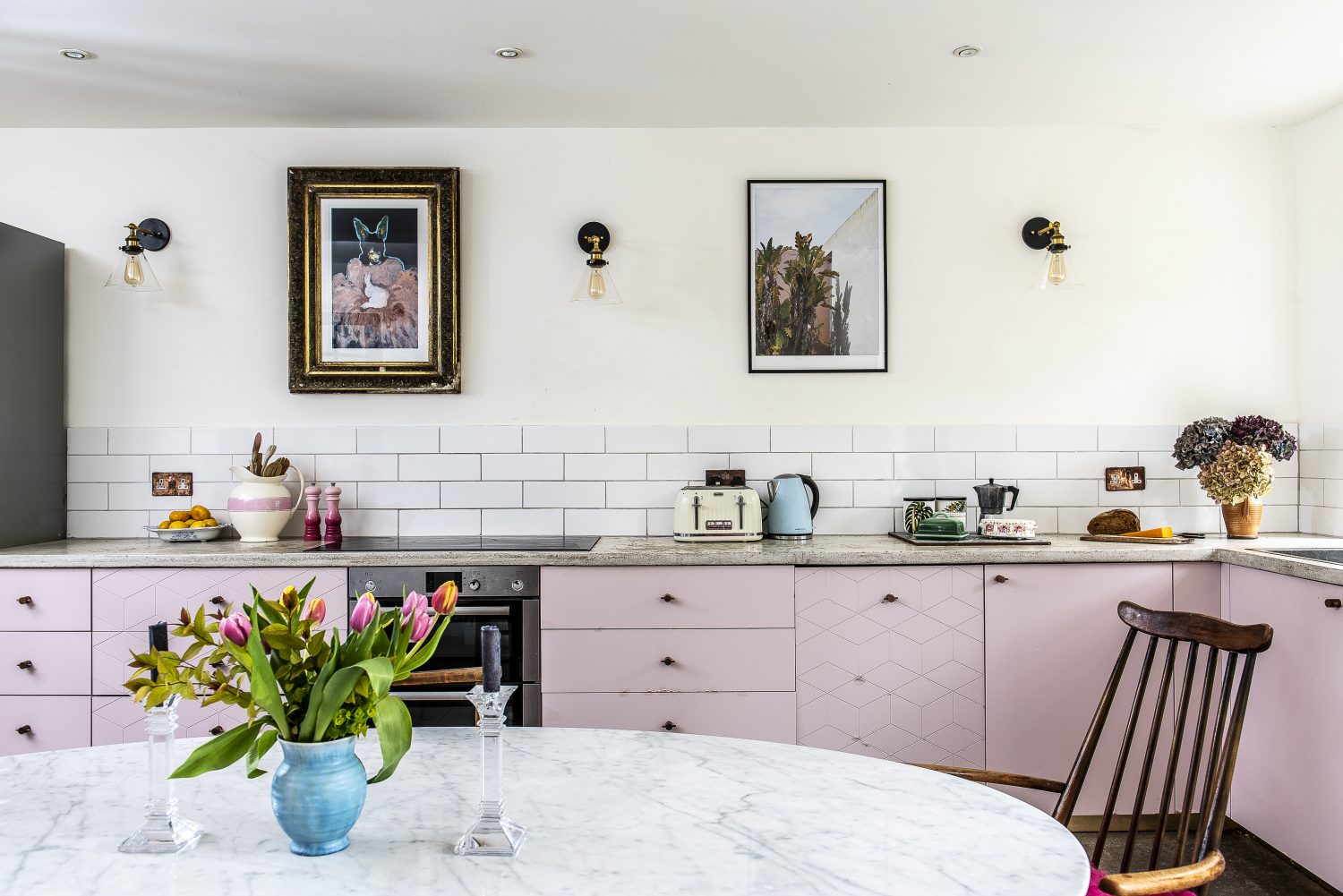 The working part of the kitchen is confined to one wall with cupboards painted a dusty pink, some with a geometric pattern in copper. The worktop is poured concrete and the wall lights are from eBay