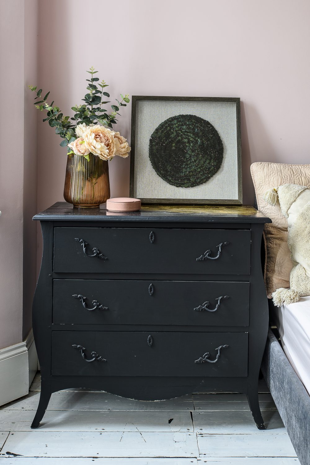 A guest bedroom has a distinct powder room feel with pale pink walls and iridescent feather artwork found in Homesense atop a black chest of drawers with a gold leaf top