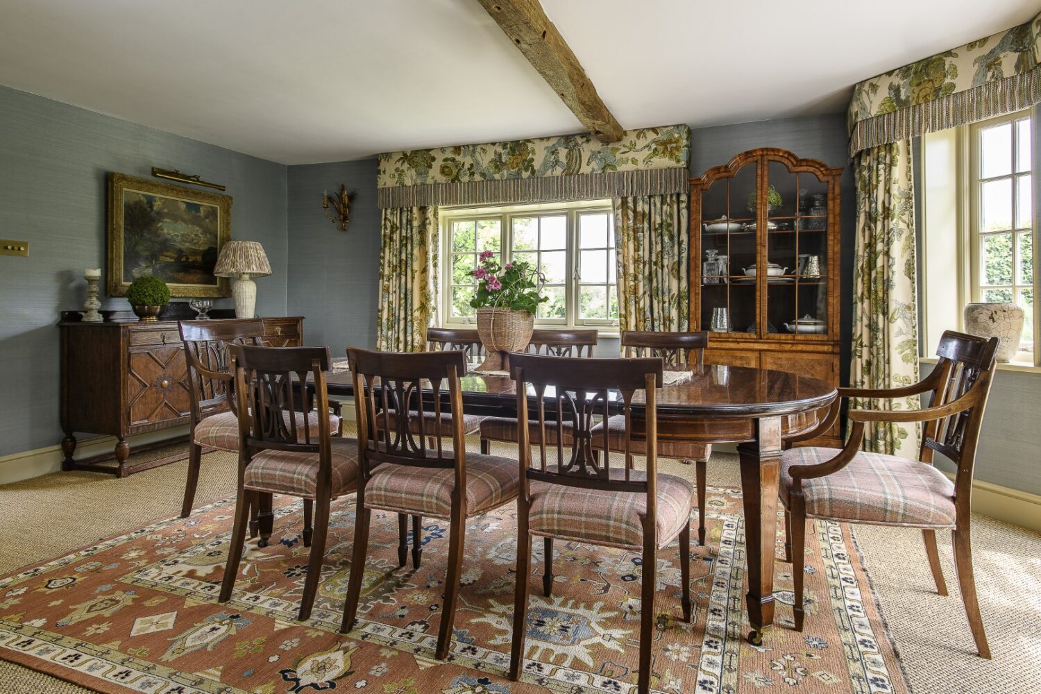 The dining room is in the older part of the house. Olya concealed the exposed pipe work with box pelmets above the curtains. An antique display cabinet has been strategically placed between two windows, and an early 19th century arts and crafts credenza sits below a beautiful piece of art. An antique rug is perfect for layering.