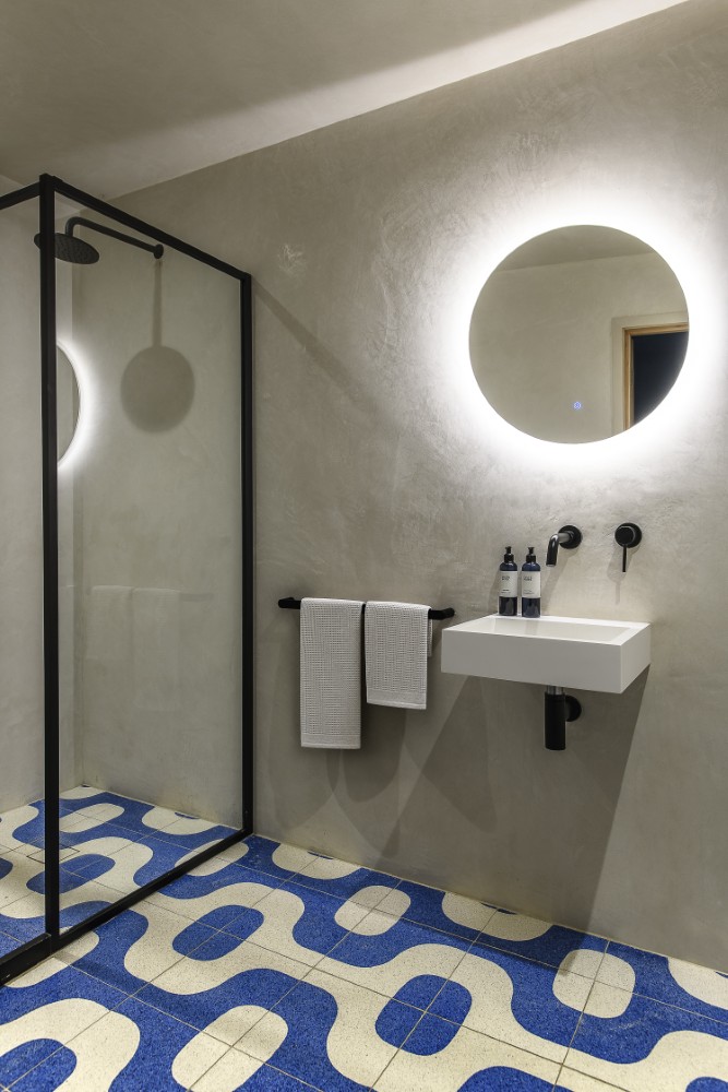 Bold blue flooring brings a playful graphic twist to the pared-back bathroom, where soft plaster walls and black-framed glazing create a striking contemporary contrast